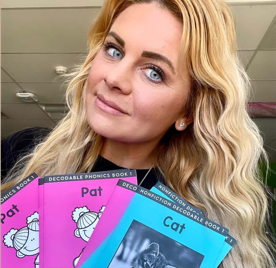 Teacher holding educational books titled 'Pat' and 'Cat' in a classroom setting